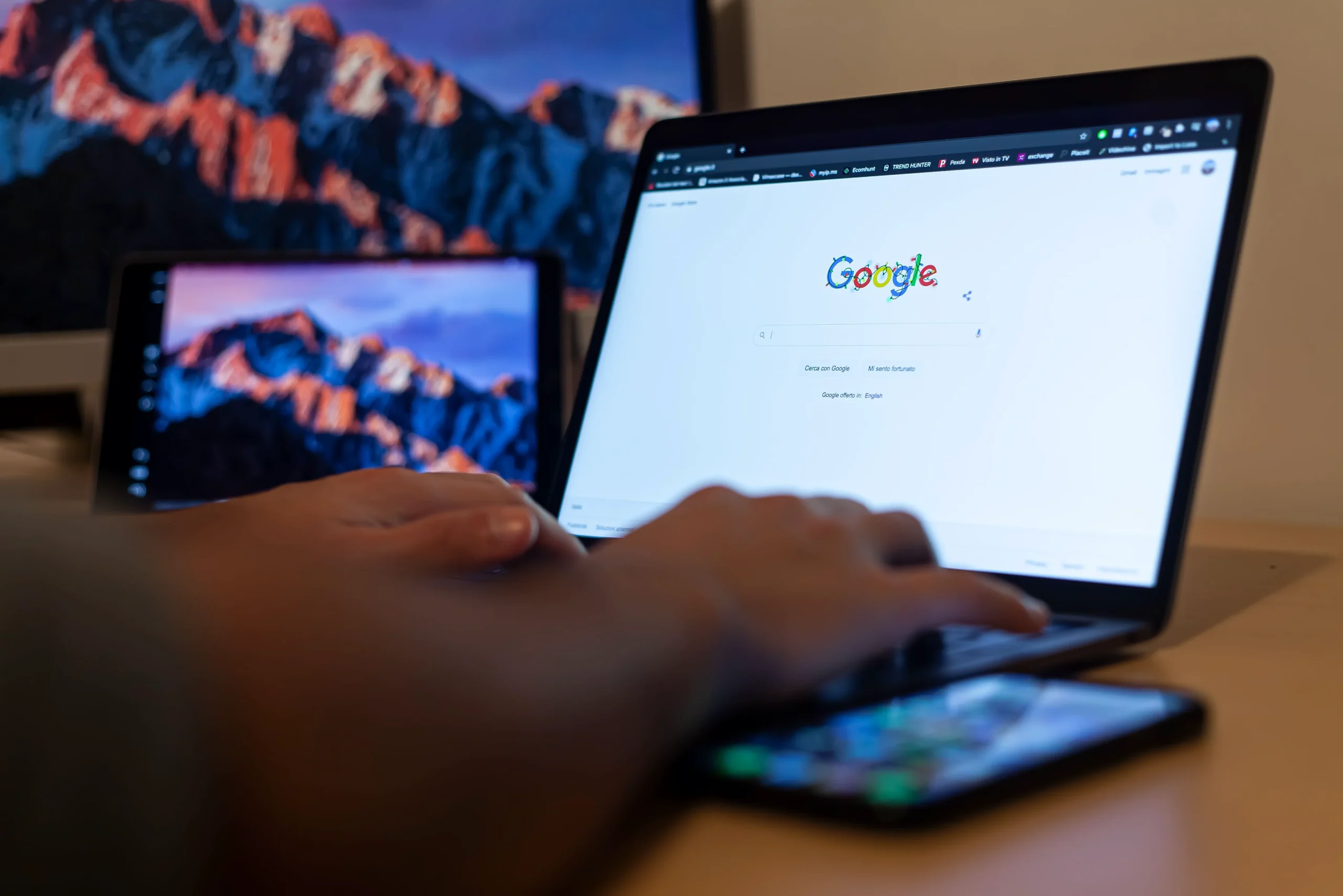 Des mains sont posées sur le clavier d’un ordinateur affichant une page de recherche Google, avec un smartphone devant et une tablette à côté.